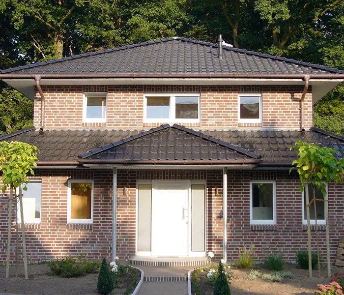 Ansicht eines zweistöckigen Backsteinhauses mit dunklem Ziegeldach und weißer Eingangstür.