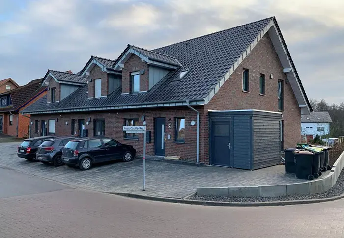 Referenz Hausbau: Mehrfamilienhaus mit dunklem Ziegeldach, roter Fassade und gepflasterter Zufahrt.