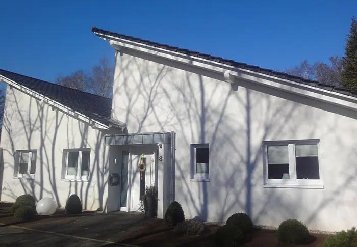Referenz Hausbau: Fassade eines modernen, weißen Einfamilienhauses mit dunklem Dach und Fenster, Baumschatten sichtbar.