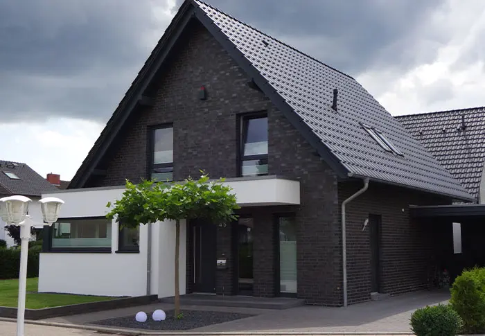 Referenz Hausbau: Modernes Einfamilienhaus mit dunklem Ziegel und Satteldach, Baum im Vorgarten.