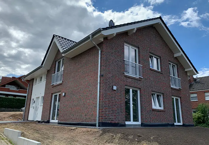 Ansicht eines zweistöckigen Hauses mit Ziegelmauerwerk, weißen Fensterrahmen und kleinem Balkon unter blauem Himmel.