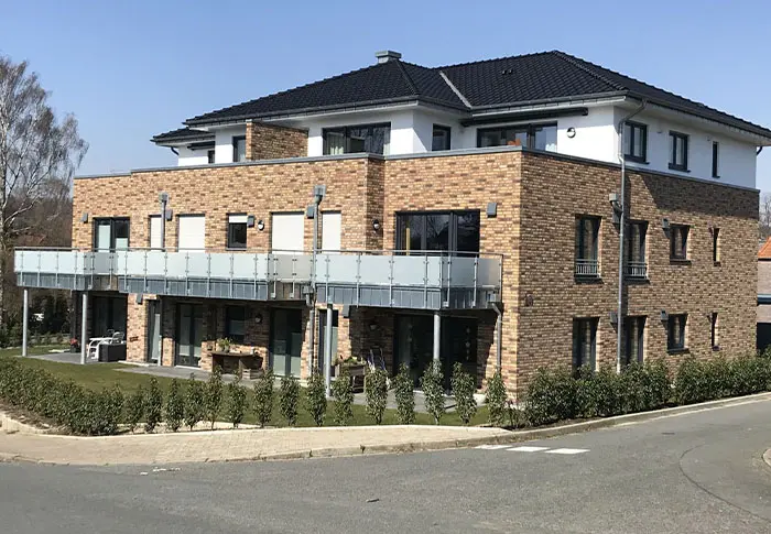 Referenz-Wohnungsbau: Mehrfamilienhaus mit Ziegelsteinfassade, Balkonen und dunklem Dach an einem sonnigen Tag.