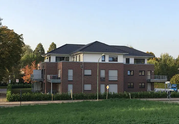 Ansicht eines modernen Mehrfamilienhauses mit Ziegelsteinfassade und dunklem Dach, umgeben von Grünflächen.
