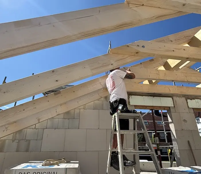 Handwerker auf einer Leiter bei der Montage von hölzernen Dachbalken auf einer Baustelle unter blauem Himmel