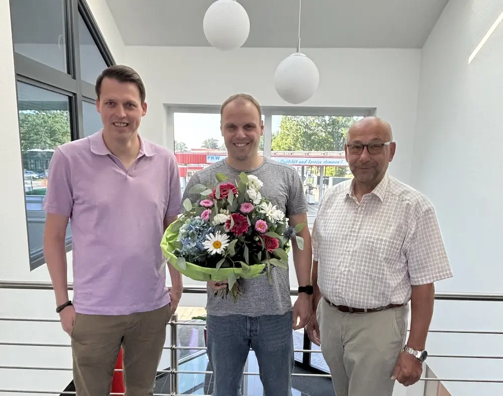 Henning Krümberg mit Blumenstrauß zwischen zwei Kollegen in einem hellen Bürogebäude von Middendorf Bau