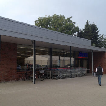 Ansicht eines Aldi-Marktes mit Einkaufswagen, Fahrrädern und einer Person vor dem Eingang.