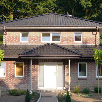 Ansicht eines zweistöckigen Backsteinhauses mit dunklem Ziegeldach und weißer Eingangstür.