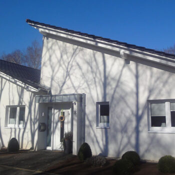 Referenz Hausbau: Fassade eines modernen, weißen Einfamilienhauses mit dunklem Dach und Fenster, Baumschatten sichtbar.