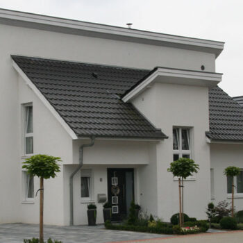 Referenz Hausbau: Modernes, weißes Einfamilienhaus mit dunklem Ziegeldach und gepflegtem Vorgarten.
