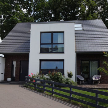 Referenz Hausbau: Modernes Einfamilienhaus mit grauen Dachziegeln, weißen Wänden und dunklen Fenstern.