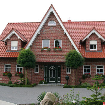 Referenz Hausbau: Fassade eines zweistöckigen Einfamilienhauses mit rotem Ziegeldach und gepflegtem Vorgarten.
