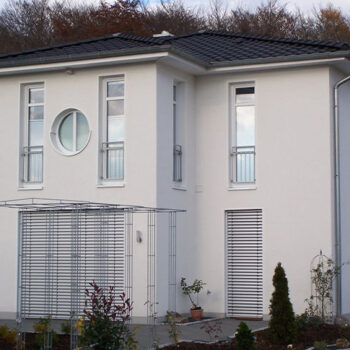 Ansicht eines modernen, weißen Hauses mit dunklem Dach und runden Fenster als Referenz Hausbau.