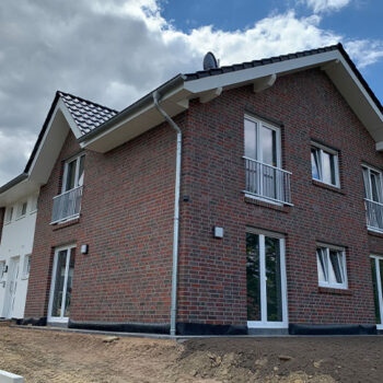 Ansicht eines zweistöckigen Hauses mit Ziegelmauerwerk, weißen Fensterrahmen und kleinem Balkon unter blauem Himmel.