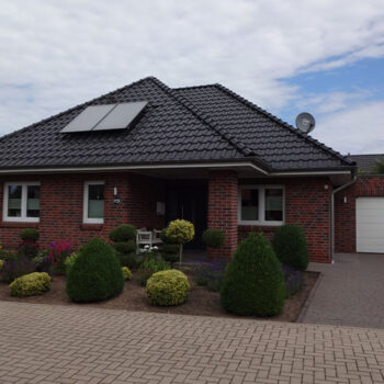 Referenz Hausbau: Ansicht eines modernen Einfamilienhauses mit roter Backsteinfassade, dunklem Ziegeldach und gepflegtem Vorgarten.