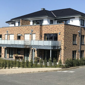 Referenz-Wohnungsbau: Mehrfamilienhaus mit Ziegelsteinfassade, Balkonen und dunklem Dach an einem sonnigen Tag.