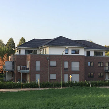 Ansicht eines modernen Mehrfamilienhauses mit Ziegelsteinfassade und dunklem Dach, umgeben von Grünflächen.